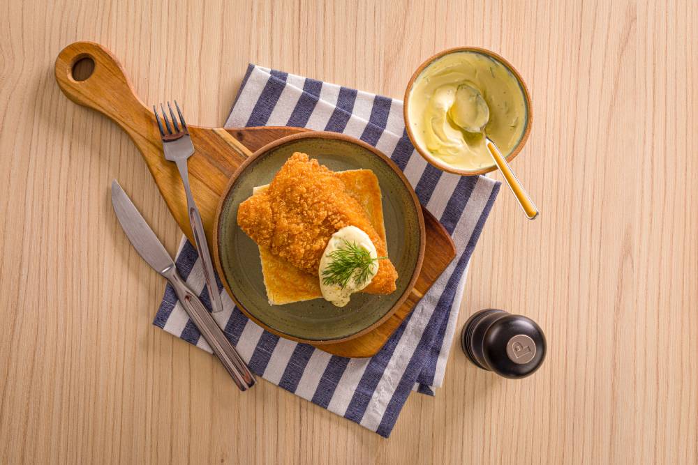 Filé de peixe empanado com molho, servido sobre uma fatia de pão de forma em uma tábua de madeira, acompanhado por um pote de molho, talheres e guardanapo listrado.
